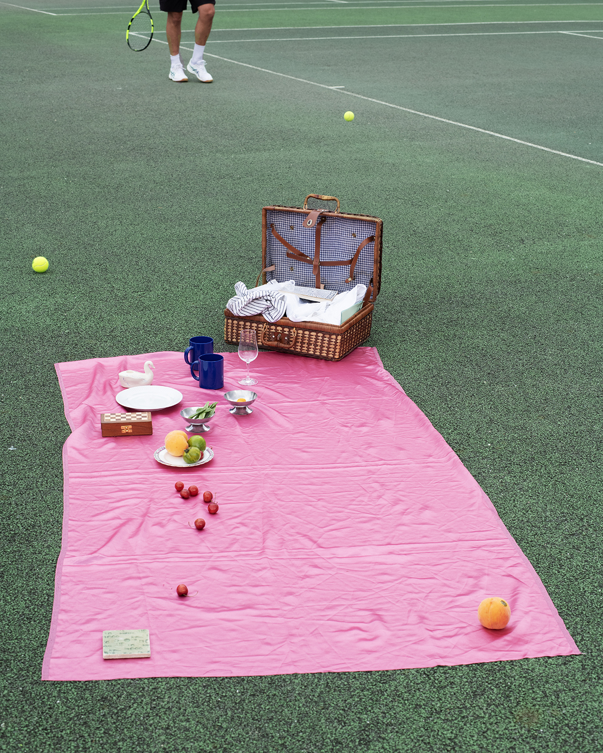 A picnic on a tennis court with a person playing tennis in the background.